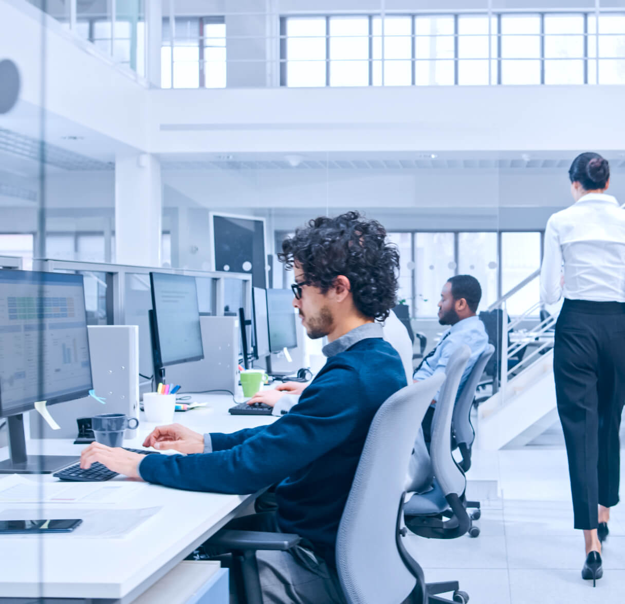 Operators working at multi-monitor workstations in a bright, modern open-plan office while a colleague walks up nearby