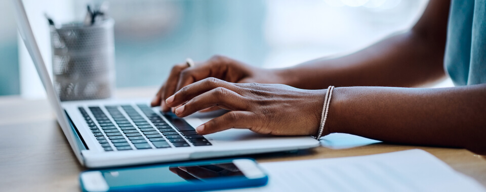 A woman's hands typing on a laptop keyboard at a desk with a smartphone and papers nearby.