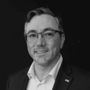 Eric Cornelius, a middle-aged man wearing glasses, a dark blazer, and an open-collared white shirt, in a black-and-white