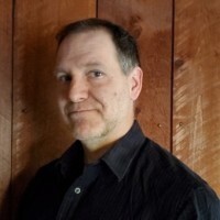 A middle-aged man with short gray hair and a goatee wearing a dark button-up shirt stands against a wooden panel wall.
