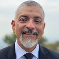 A smiling middle-aged man with gray hair and a goatee wearing a dark suit and tie in an outdoor setting.